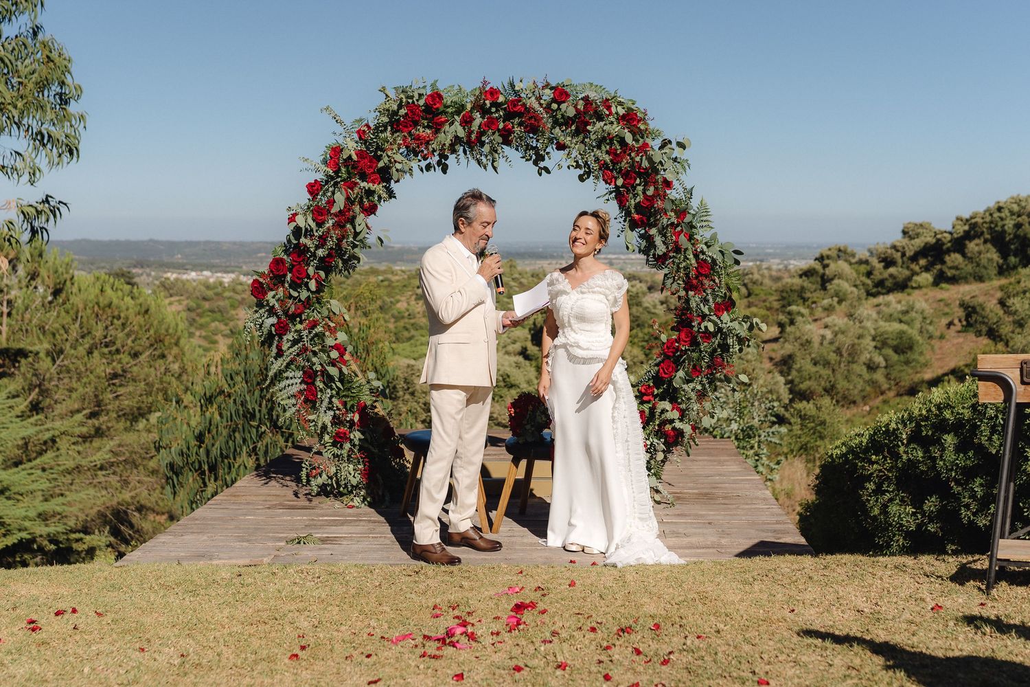 Couple exchanges vows beneath a dramatic red rose arch with panoramic views of rolling hills.