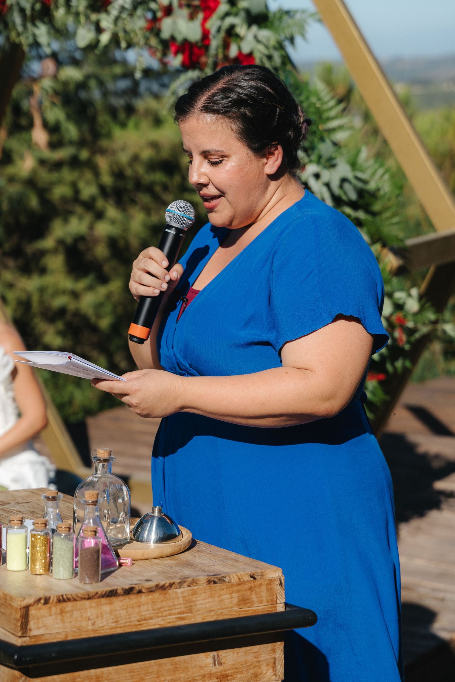 Woman in blue dress speaking into microphone at outdoor event with wooden table.