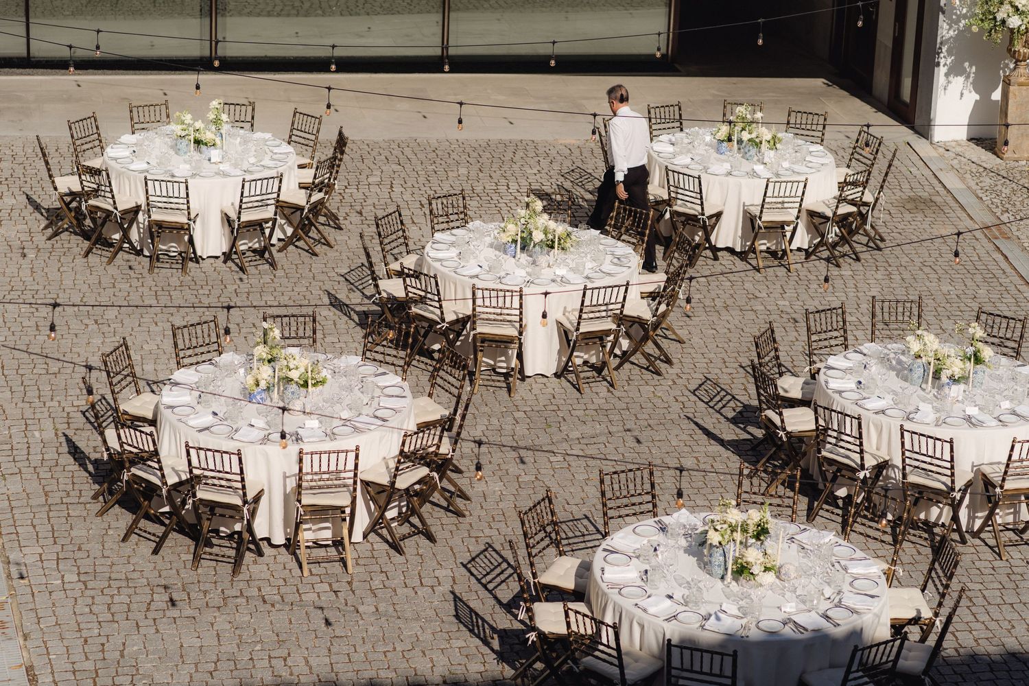 Outdoor wedding reception setup with round tables and chairs arranged on stone paving.