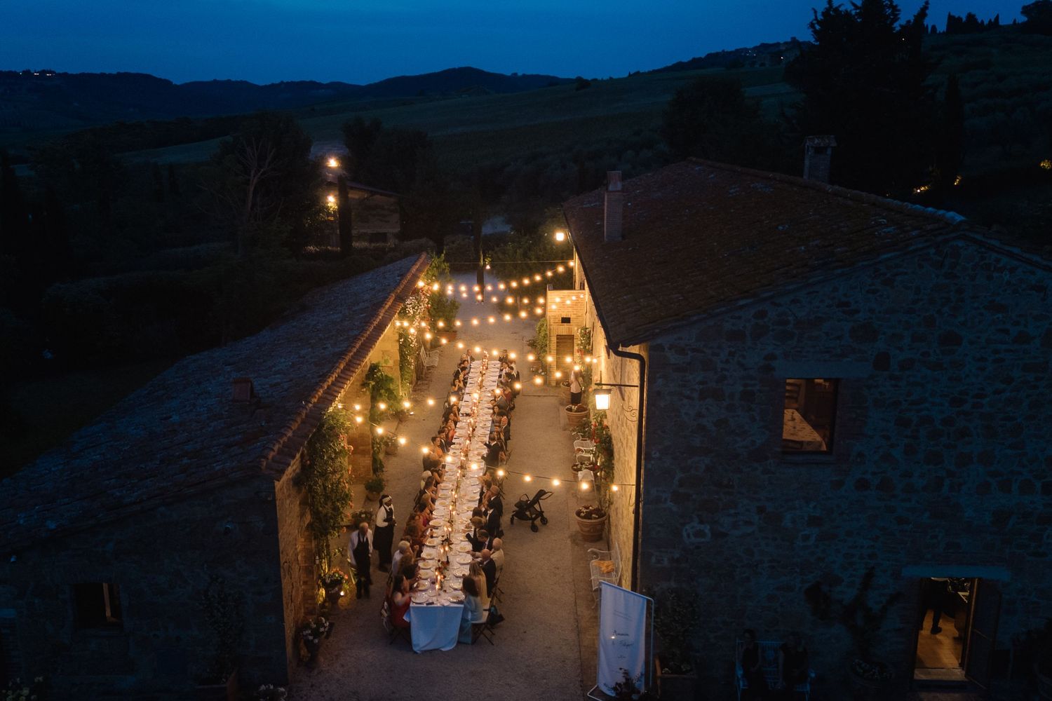 Aerial view of an elegant outdoor wedding reception with string lights and long tables at a rustic Tuscan villa at dusk.