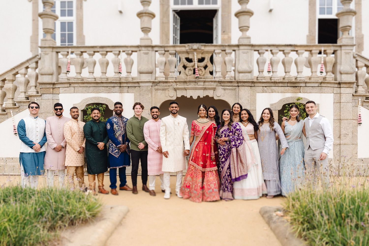 Large wedding party group photo in front of elegant building with traditional Indian formal wear.