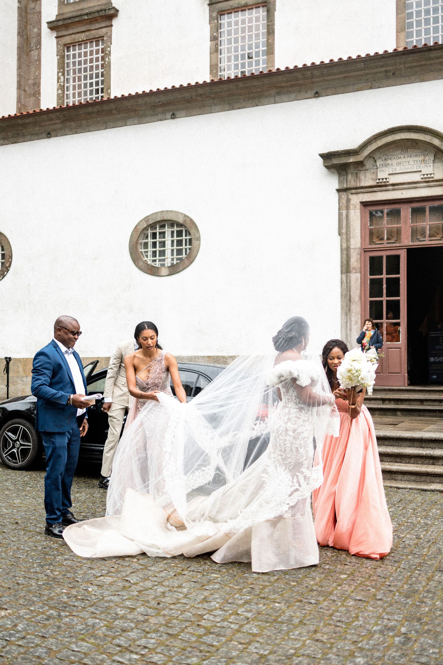 Wedding party helping with bride's veil outside historic white church building.