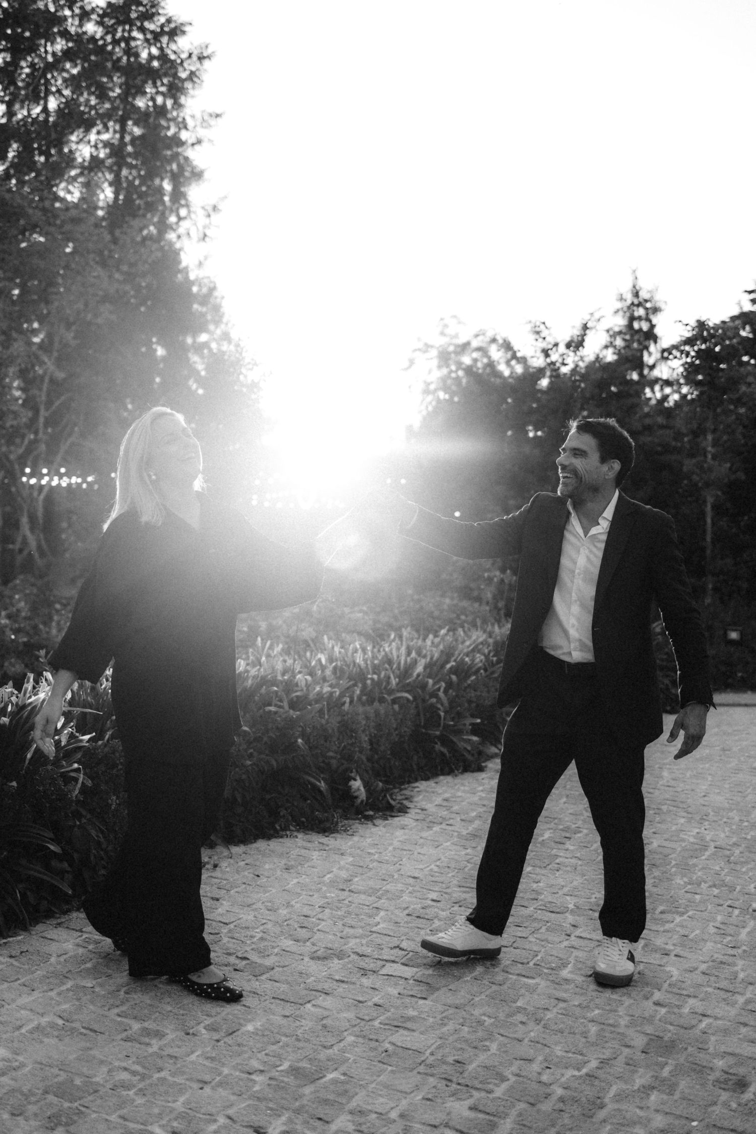 Black and white photo of a couple holding hands in a sunlit garden path, dressed in elegant formal attire.