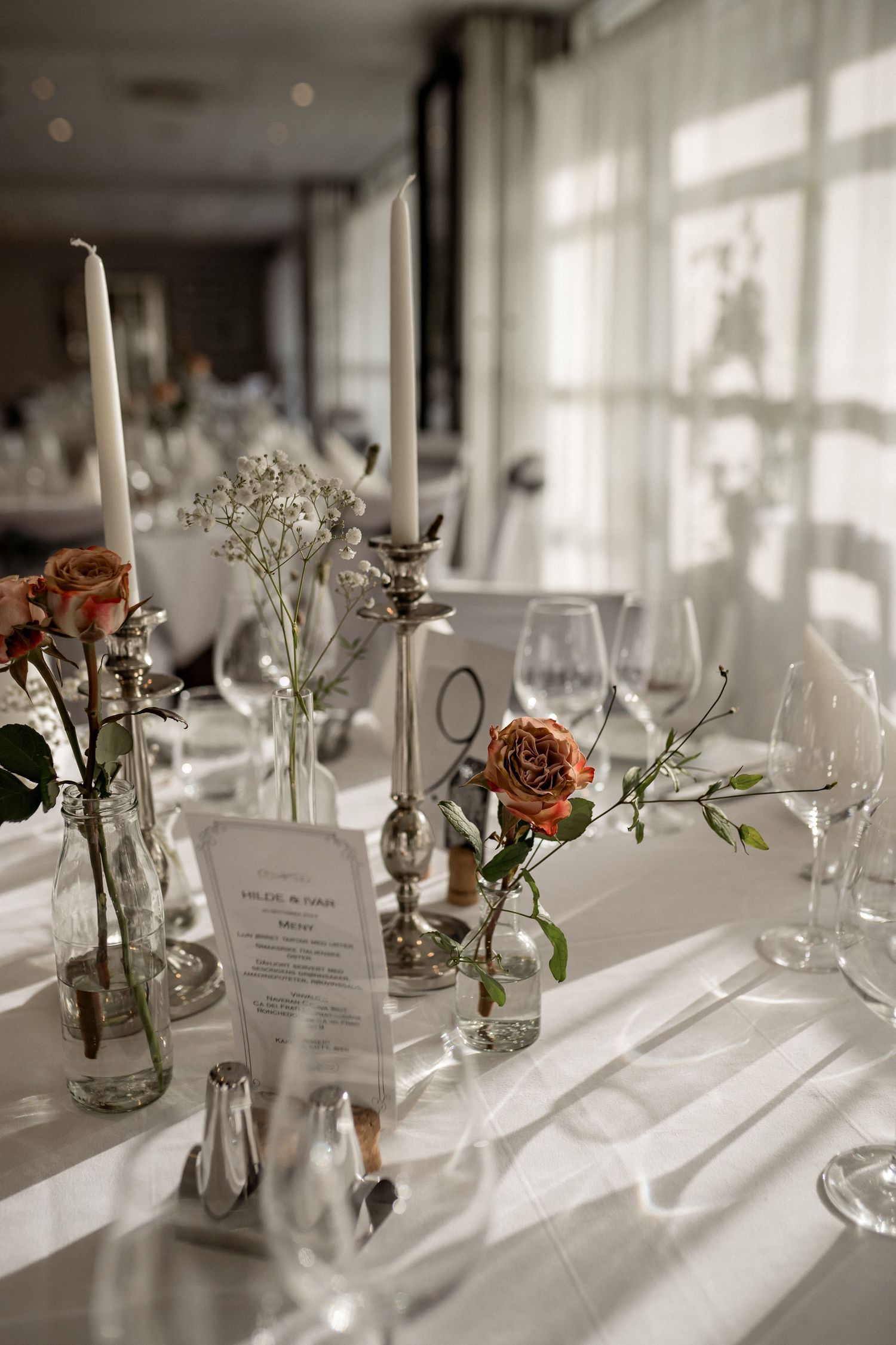 Elegant wedding table settings with delicate floral centerpieces and menus in glass vases catching sunlight.