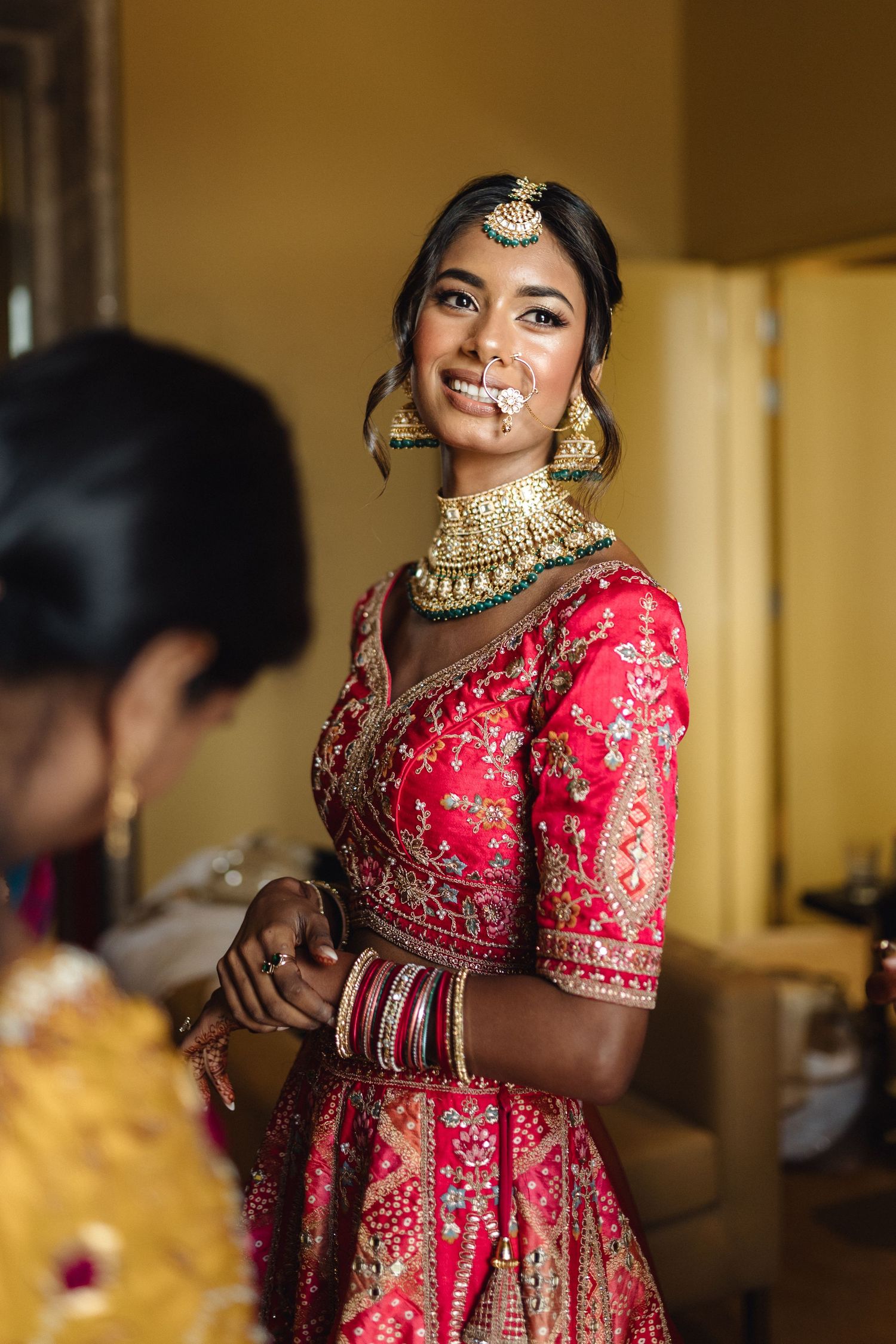 Bride in red embroidered lehenga and gold choker necklace getting ready for Indian wedding ceremony.