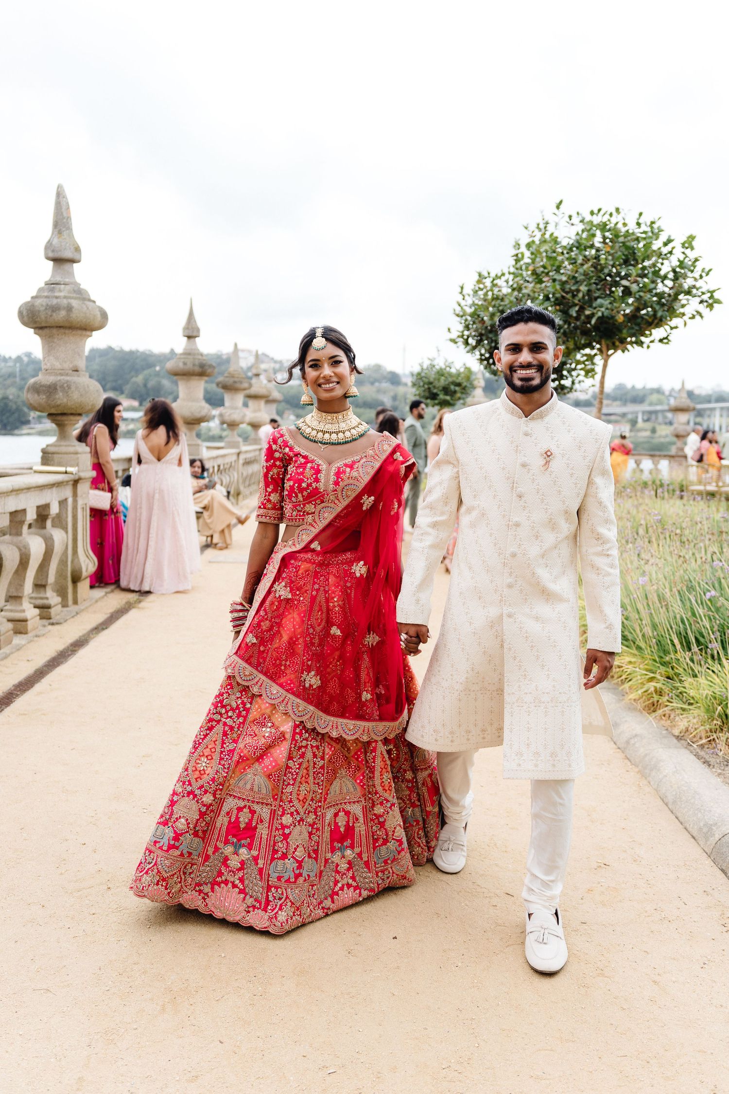 Traditional Indian wedding attire featuring red lehenga and cream sherwani on a scenic terrace.