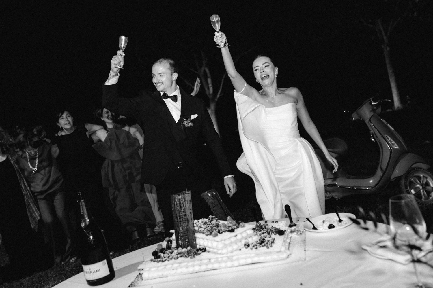 Bride and groom celebrate joyfully raising champagne glasses while cutting wedding cake at outdoor evening reception.