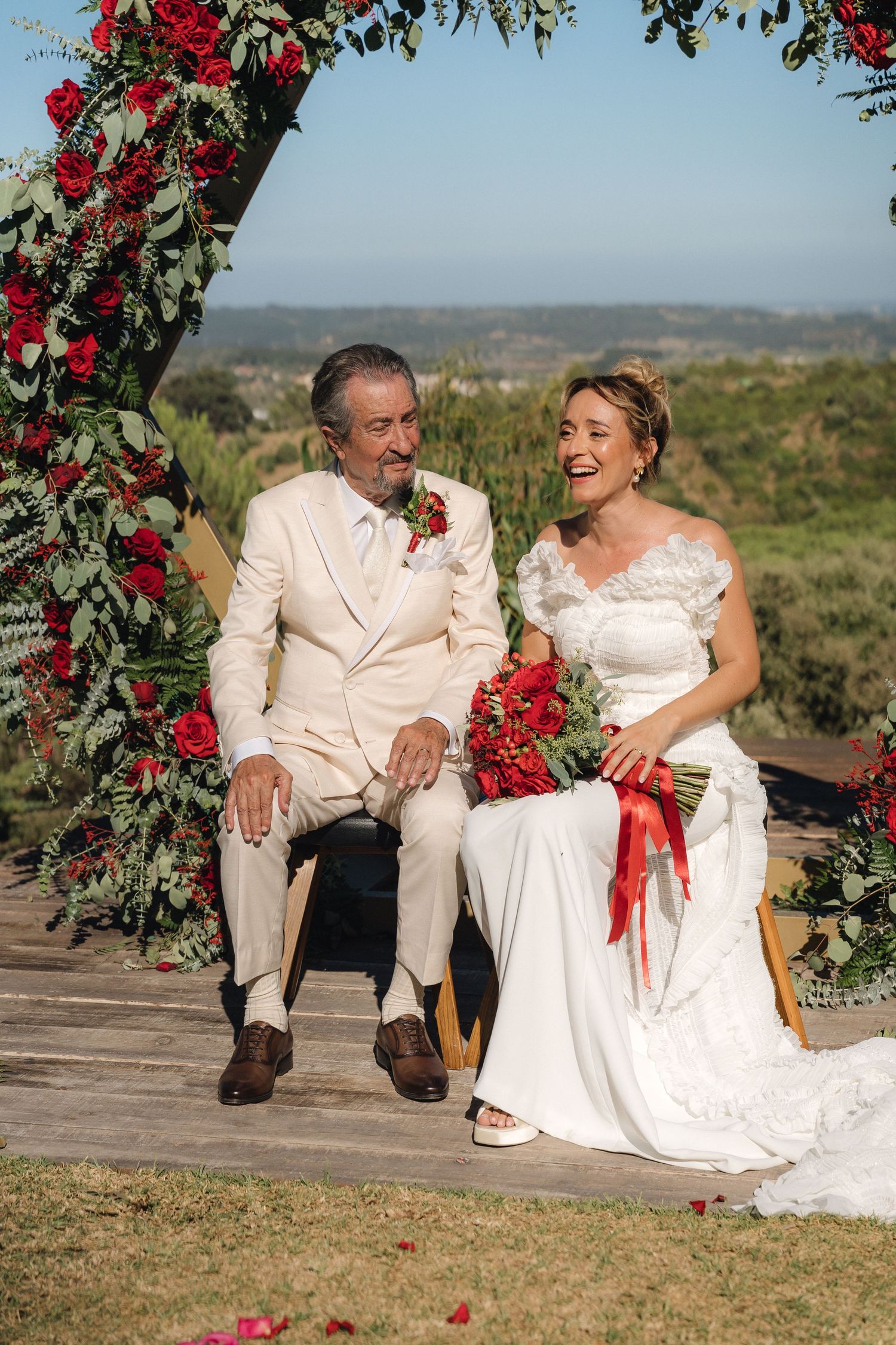 Couple sits together on wooden platform under floral arch with red roses.