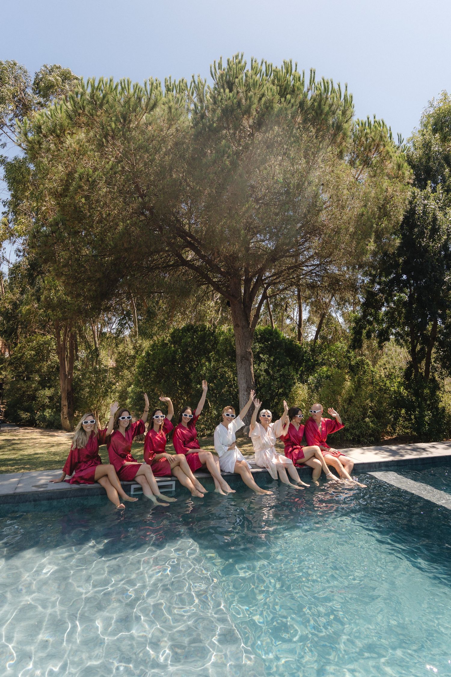 Wedding party sitting at the edge of an infinity pool with trees in the background.