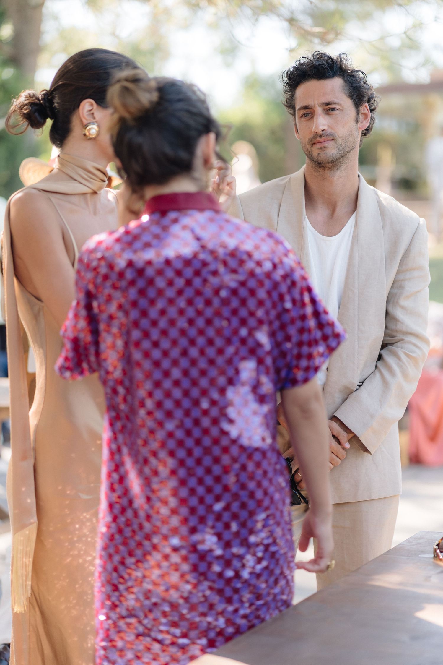 Wedding guests in formal attire interact during an outdoor celebration on a sunny day.