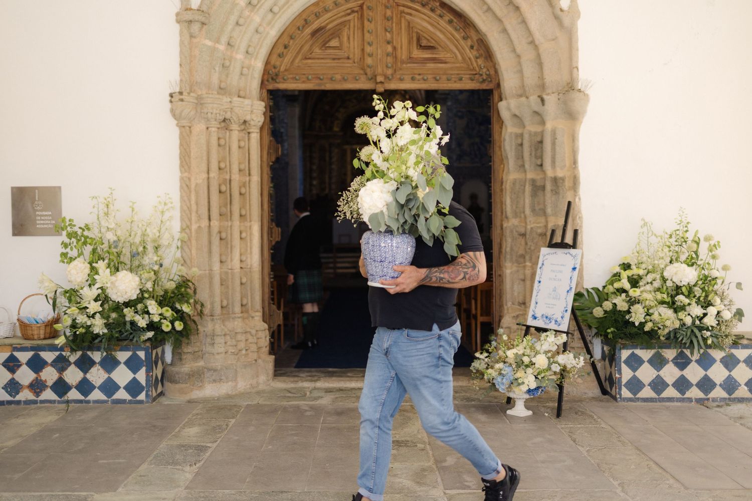 Person carrying white floral arrangement through ornate stone archway.