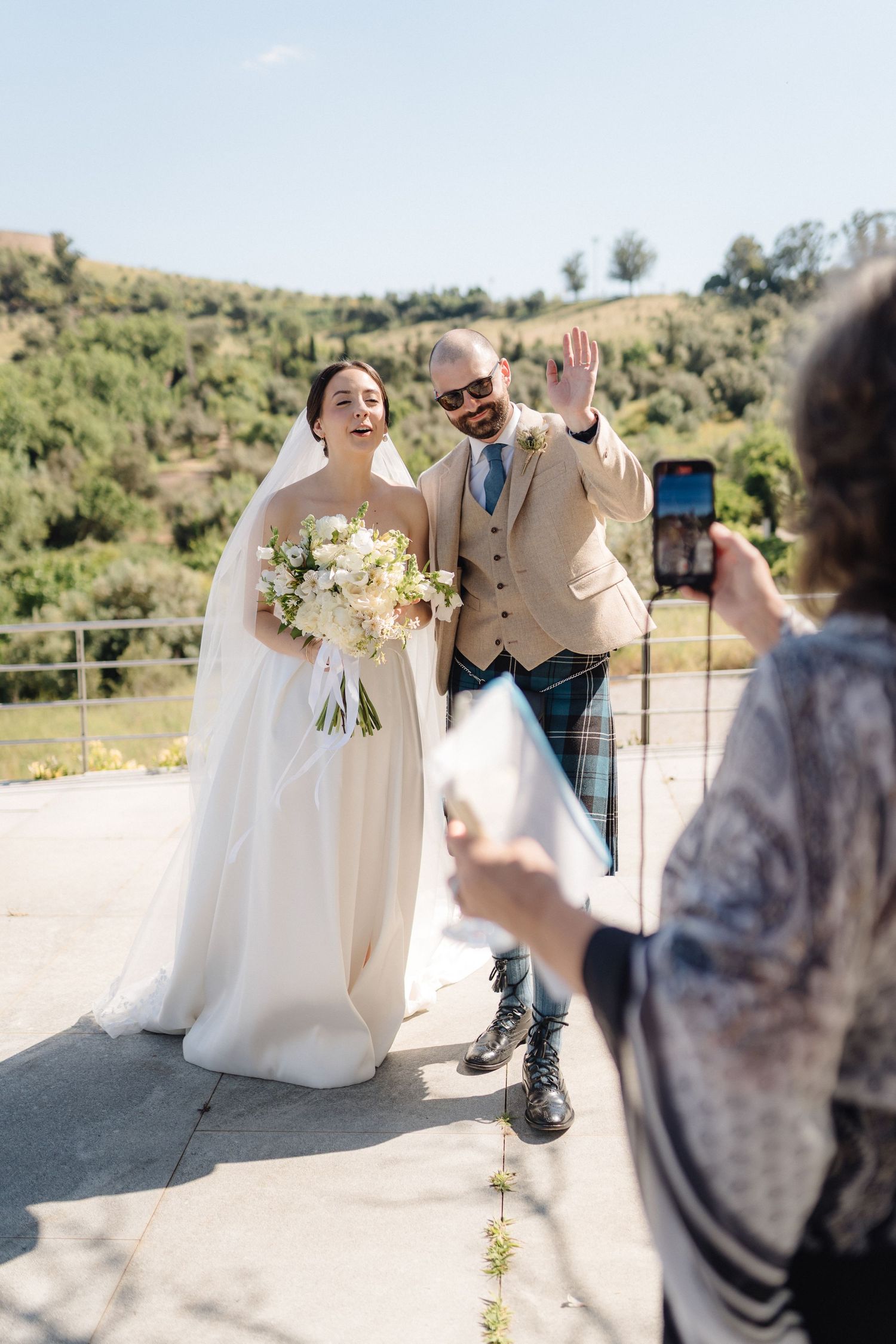 A joyful wedding moment captured by a photographer on an outdoor terrace with hills in background.