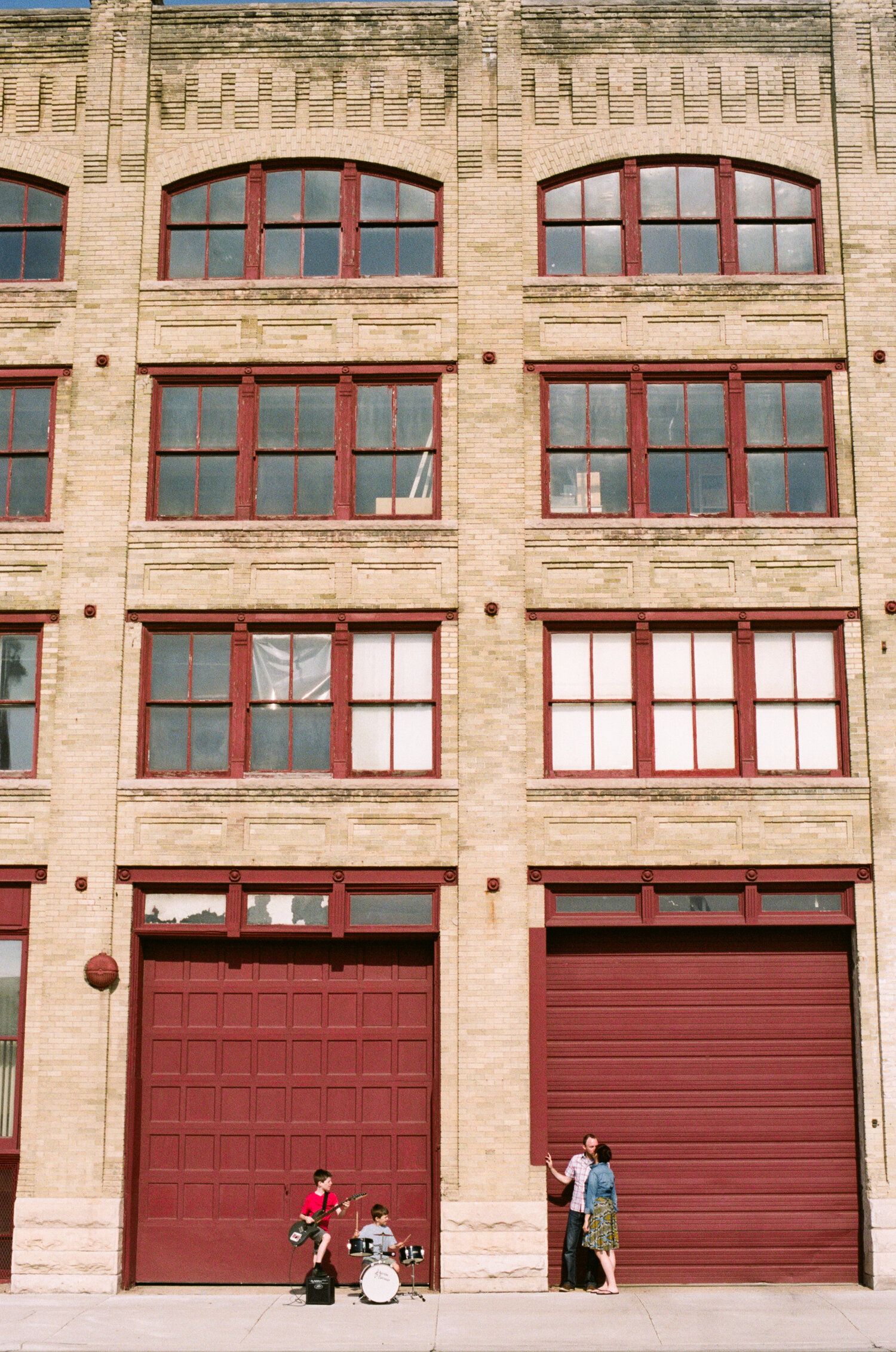 Musicians perform in front of a vintage brick building with large red garage doors and arched windows.