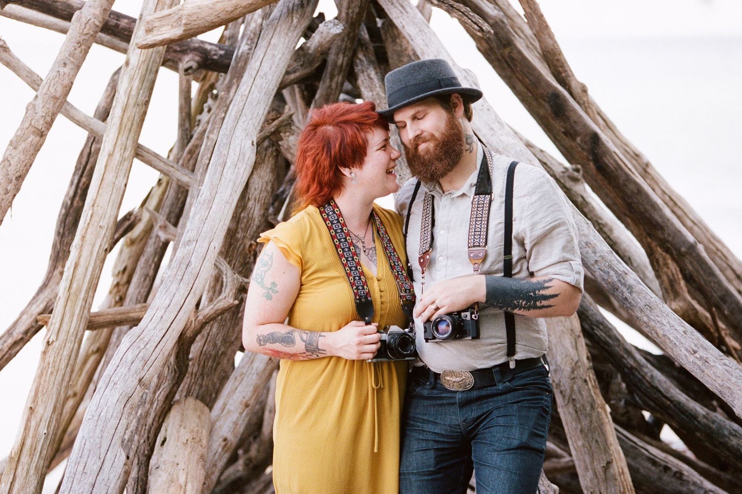 Vintage-styled couple with cameras posing by wooden structure outdoors.