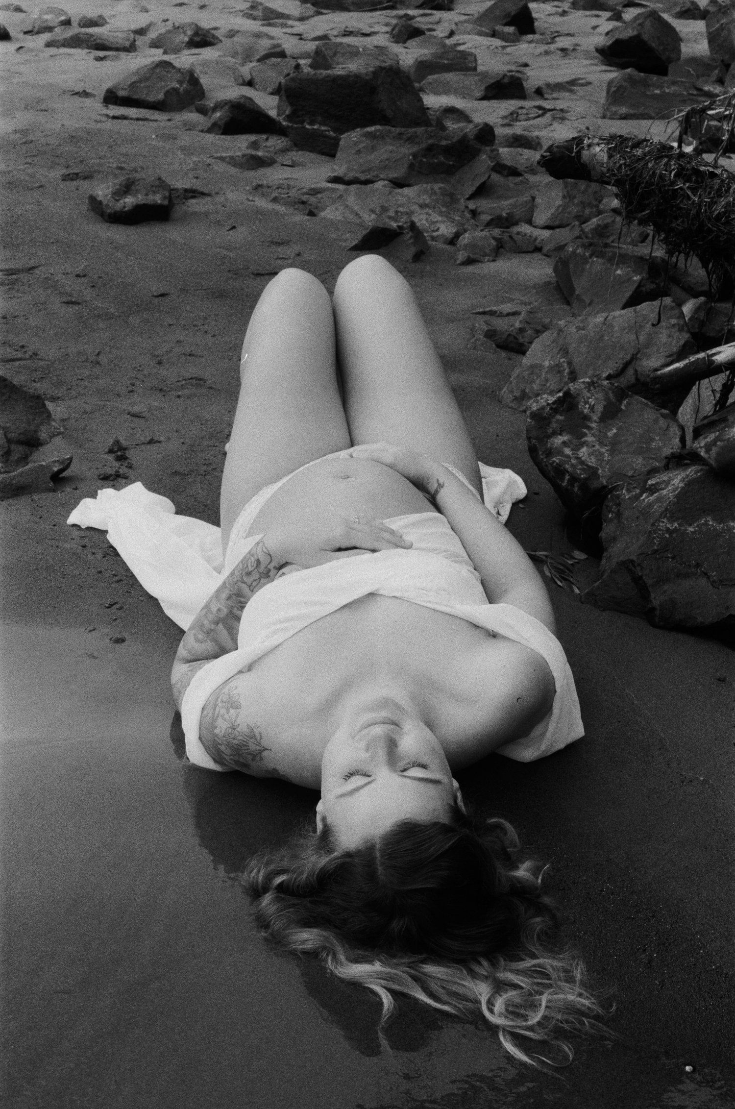 A black and white portrait of someone lying on a rocky beach in flowing white fabric.