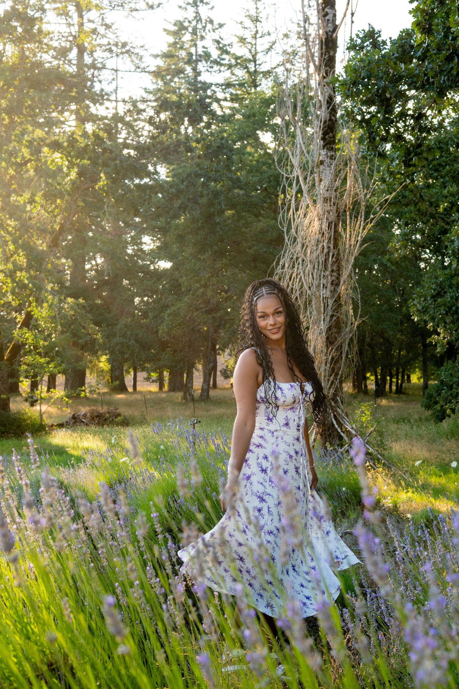 A floral sundress among blooming lavender flowers in a garden at golden hour with sunlit trees in the background.