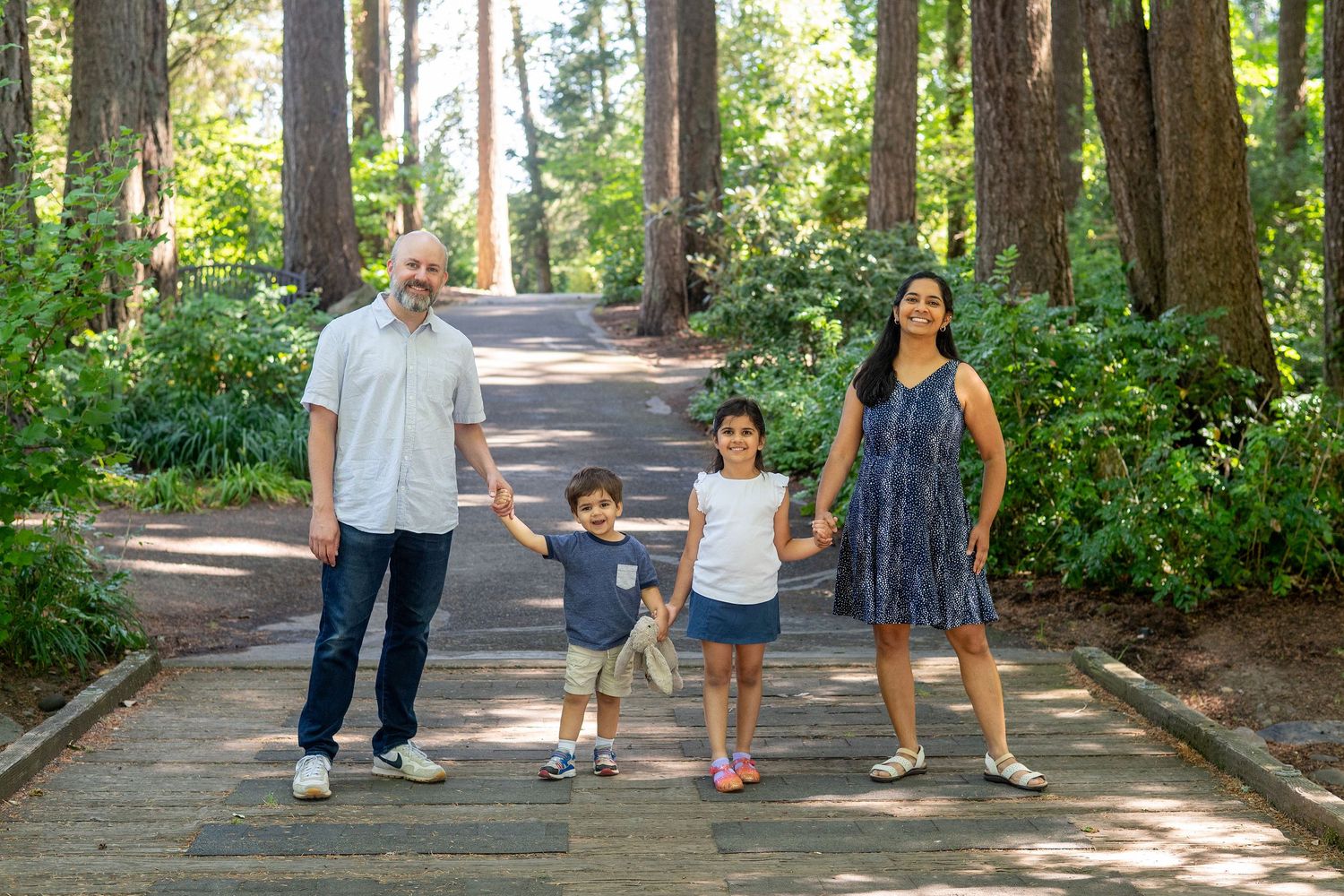 A family enjoying a sunny walk together on a tree-lined wooden boardwalk path through a lush forest.