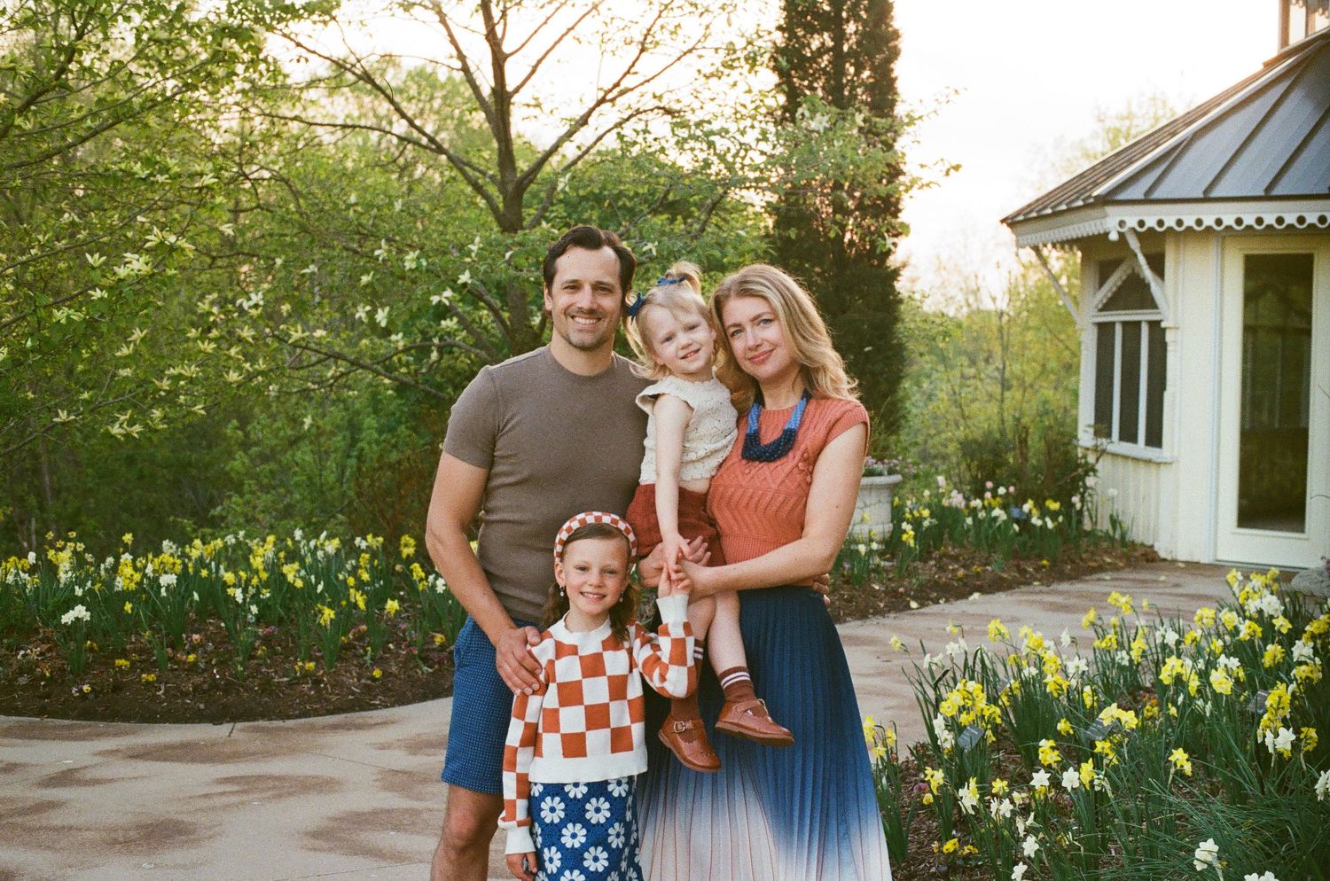 Family smiling together in spring garden with white house and yellow flowers.