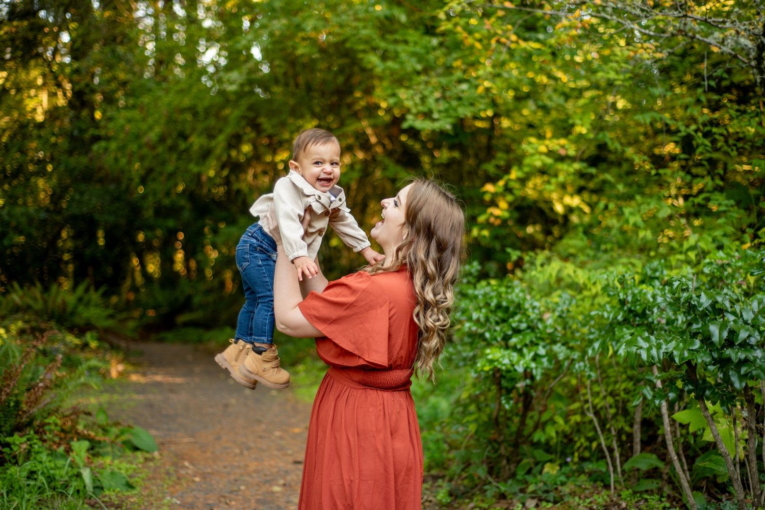 A parent in a rust-colored dress lifts a child in denim along a wooded trail during autumn.