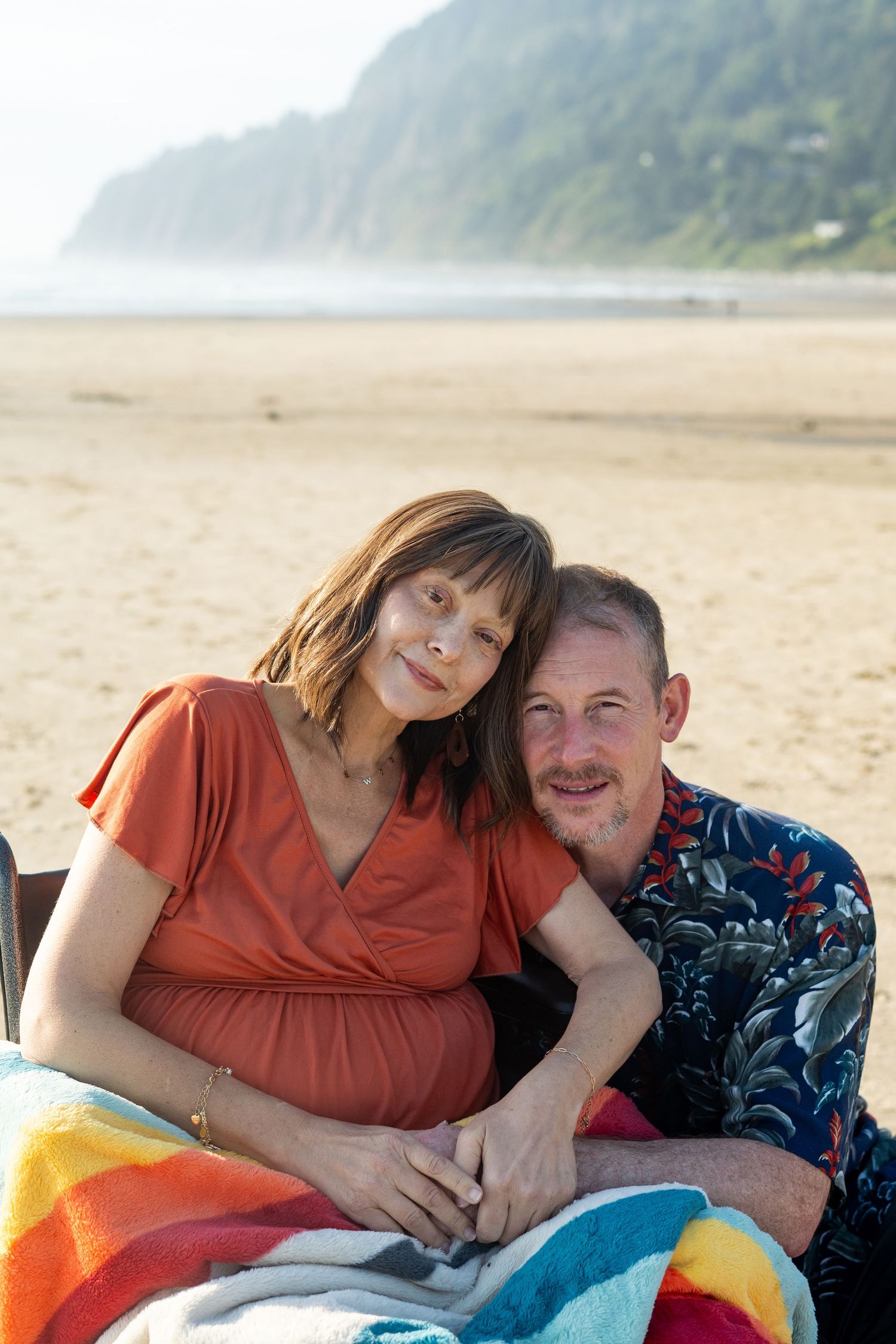 A couple rests together on colorful towels at a scenic beach with forested hills in the background.