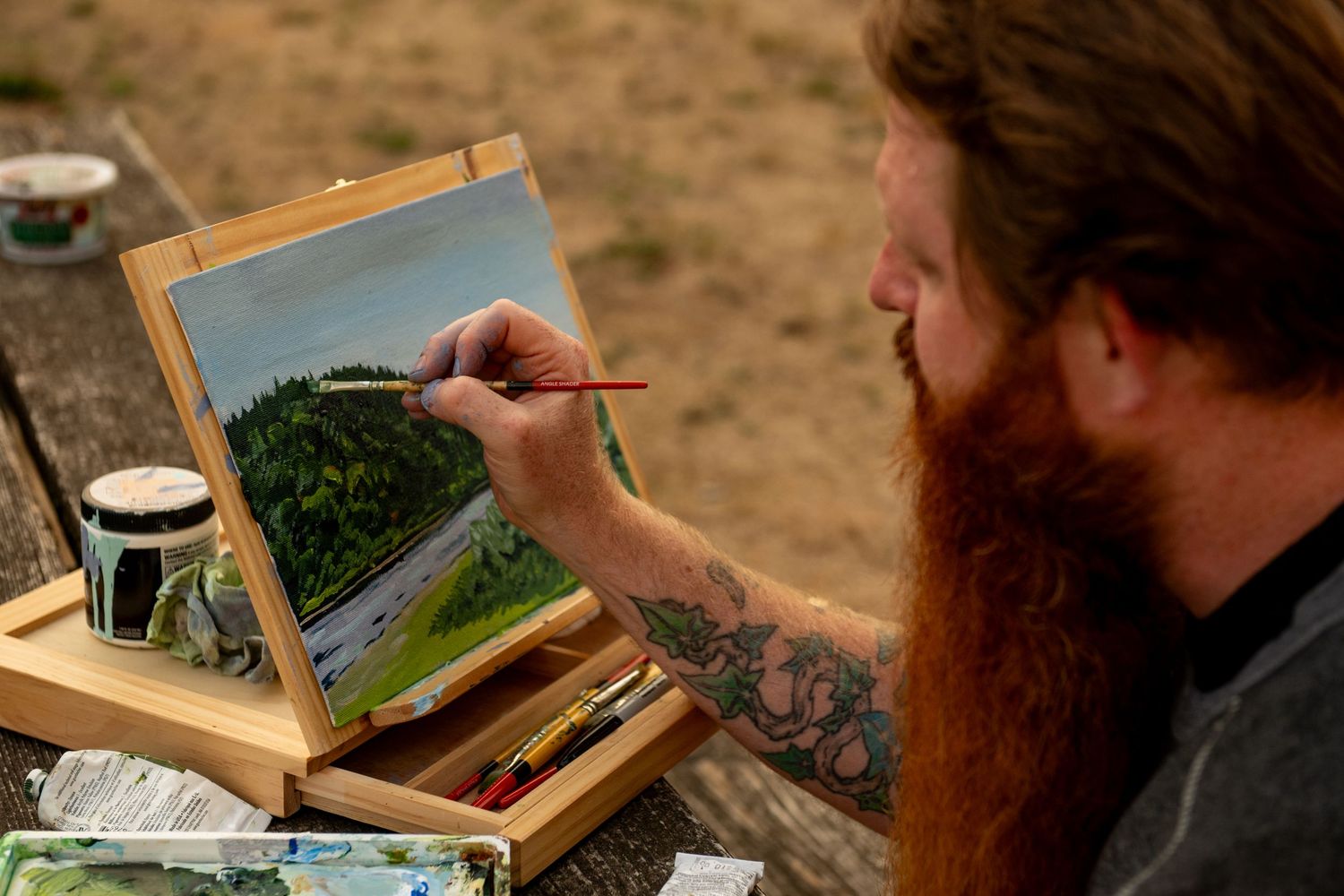An artist paints a desert landscape on a small canvas while sitting outdoors with painting supplies nearby.