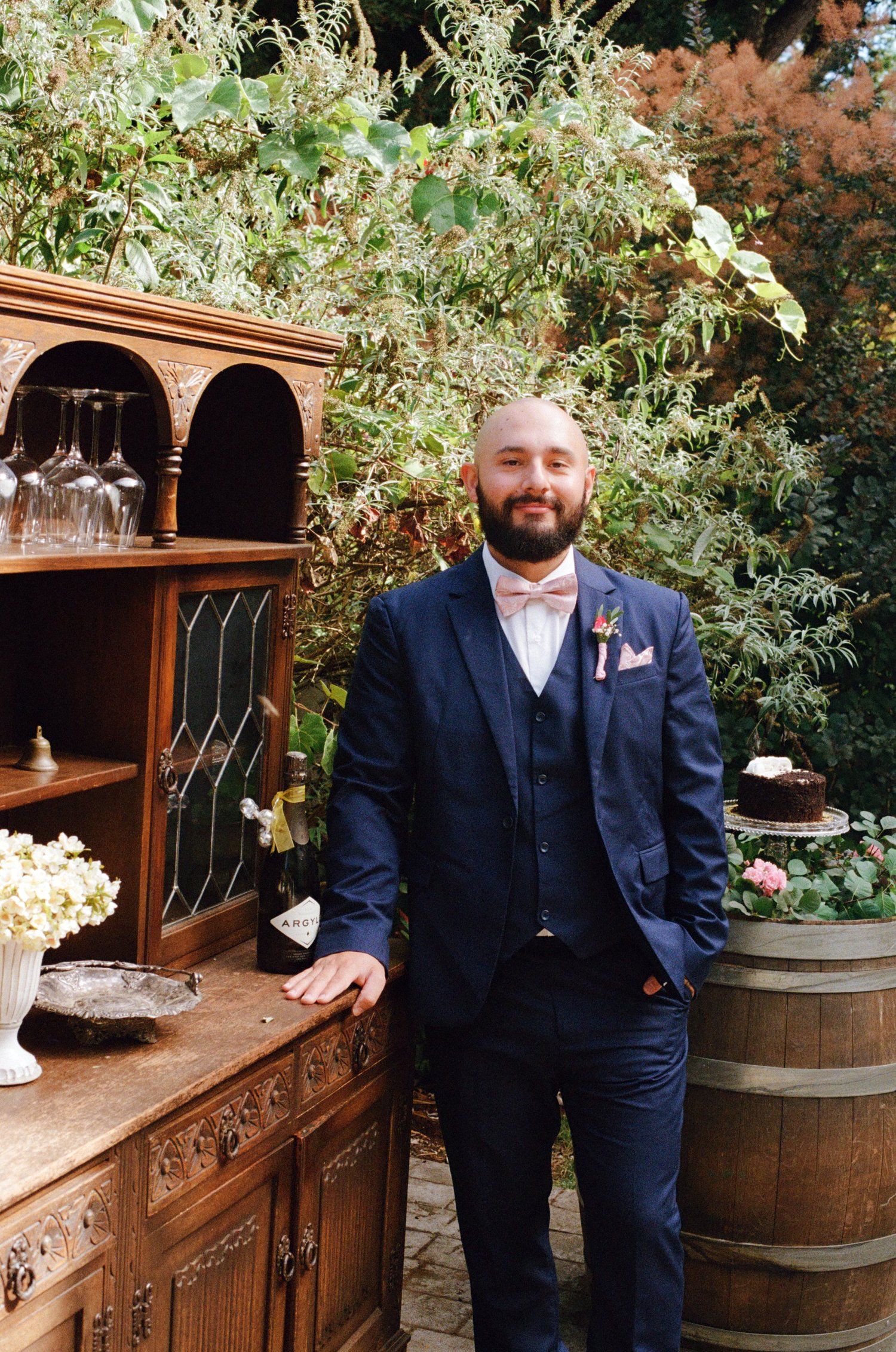 Wedding party member in navy suit poses next to rustic wooden bar with wine barrels.