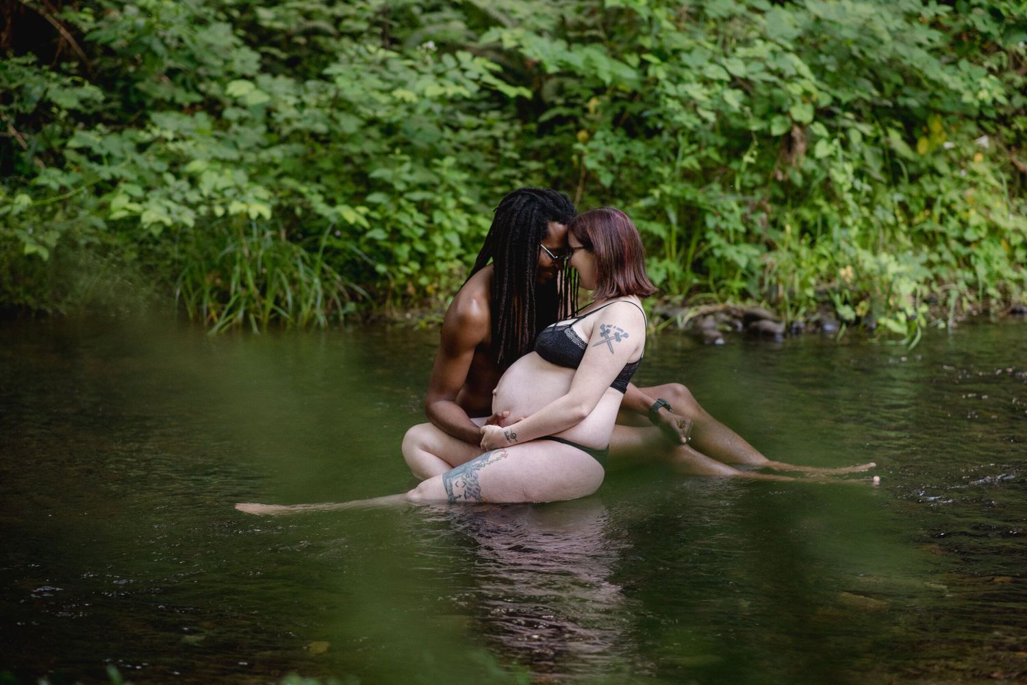 A couple embrace while sitting in shallow creek waters surrounded by lush green foliage on a summer day.