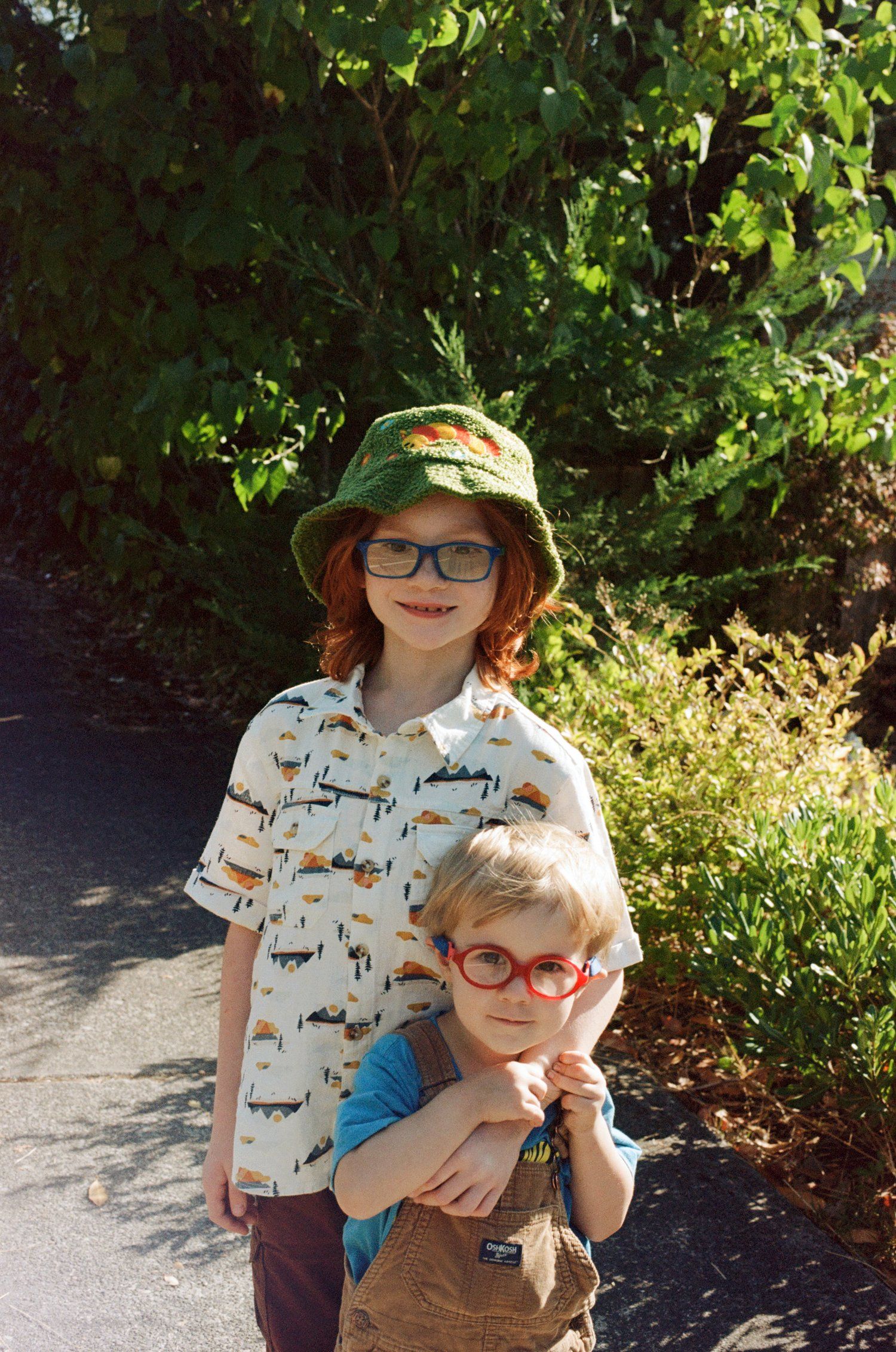 Two young people wearing glasses and sun hats pose together on a garden path.