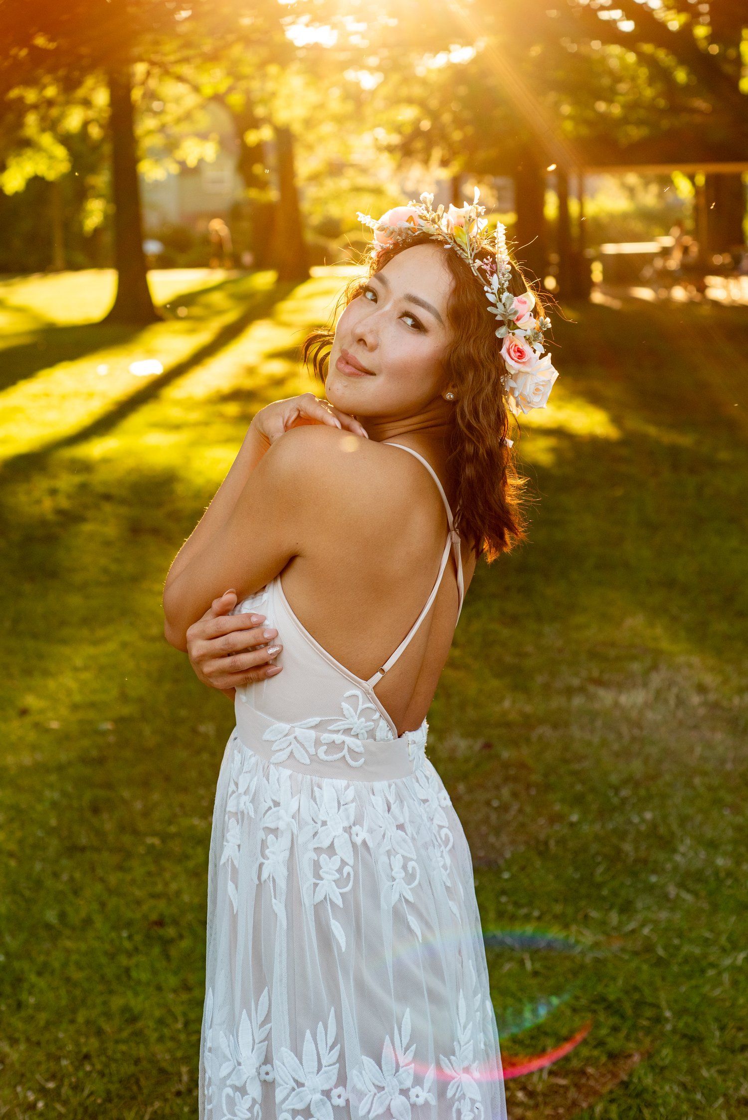 A woman in a white lace dress and floral crown poses in golden sunlit garden during sunset.