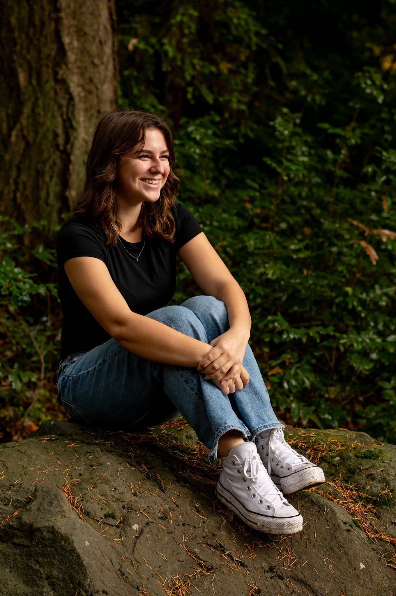 Person in casual wear sitting on rocky outcrop in forest wearing white sneakers.