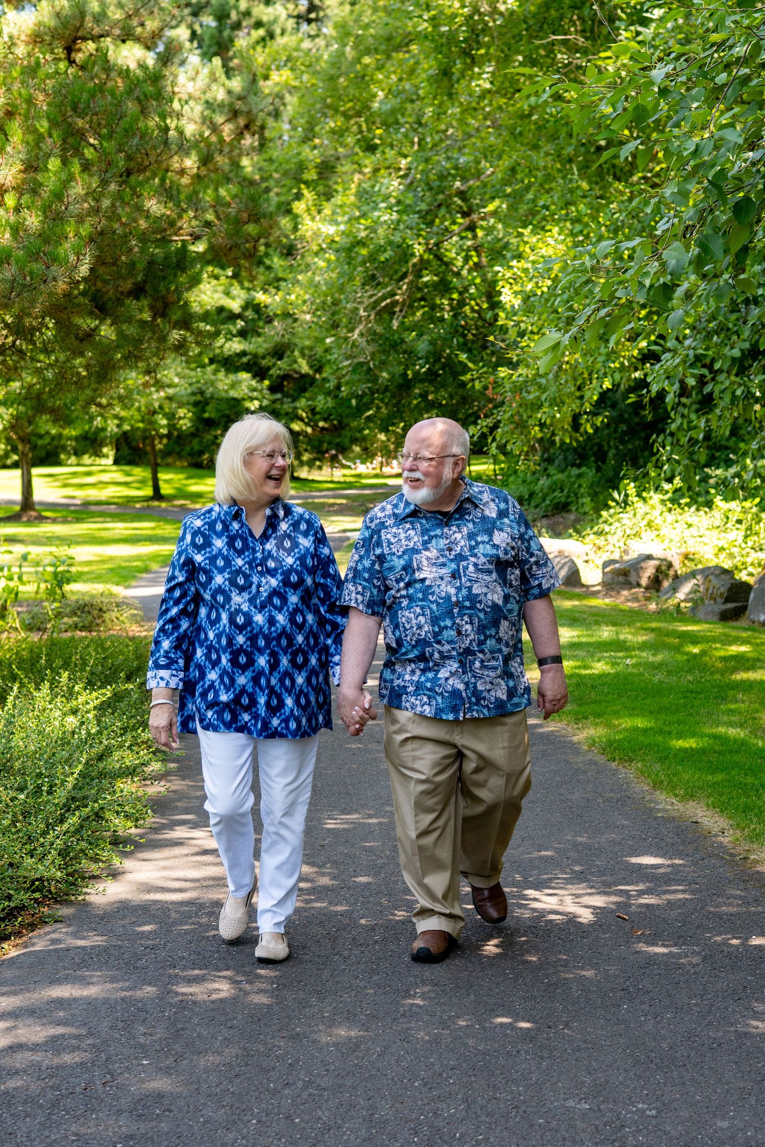 Elderly couple walking together along a tree-lined path in a sunny park while wearing coordinating blue patterned shirts.