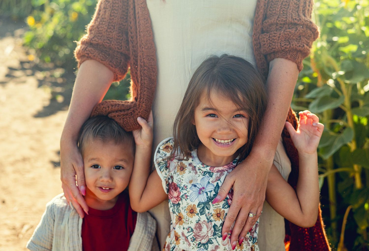 Two children in casual attire smile while standing outdoors in sunlit garden with sunflowers.