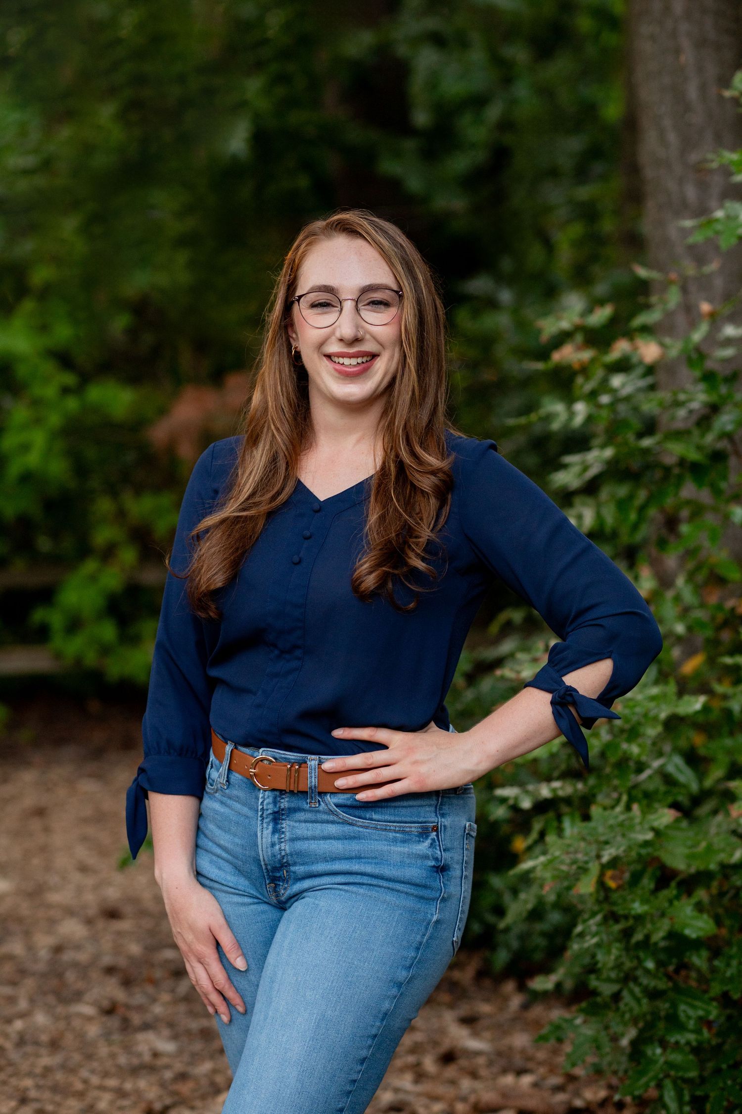 A person in a navy blue top and jeans poses casually outdoors against a green foliage background.