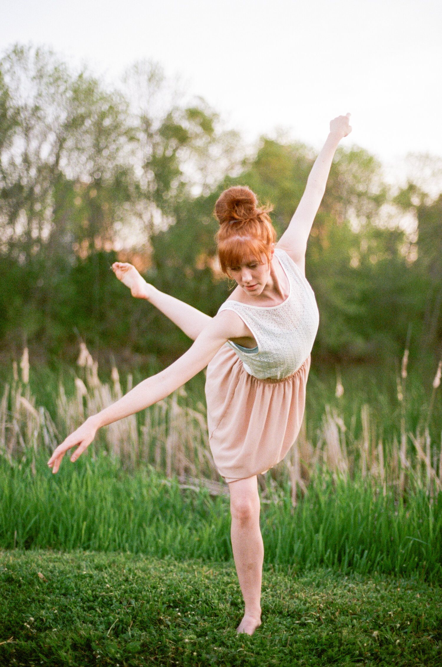 A ballerina in a pink skirt and white top performs an elegant dance pose in a grassy outdoor setting at sunset.
