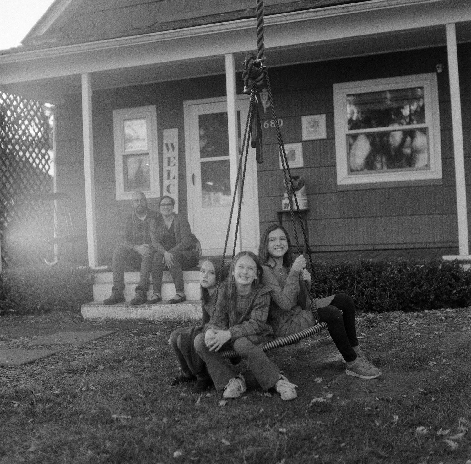 A group sits together on a porch swing in front of a classic American house with lattice windows.