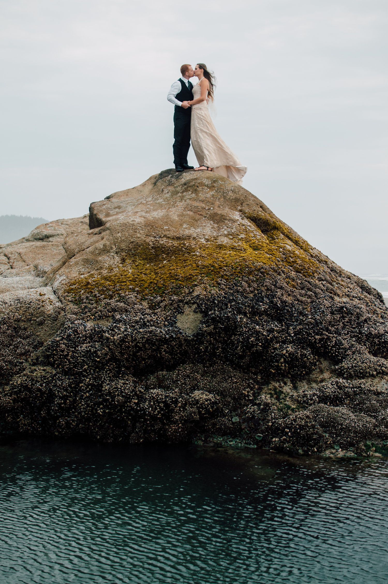 A couple stands on a rocky oceanside cliff in formal attire embracing against cloudy sky.