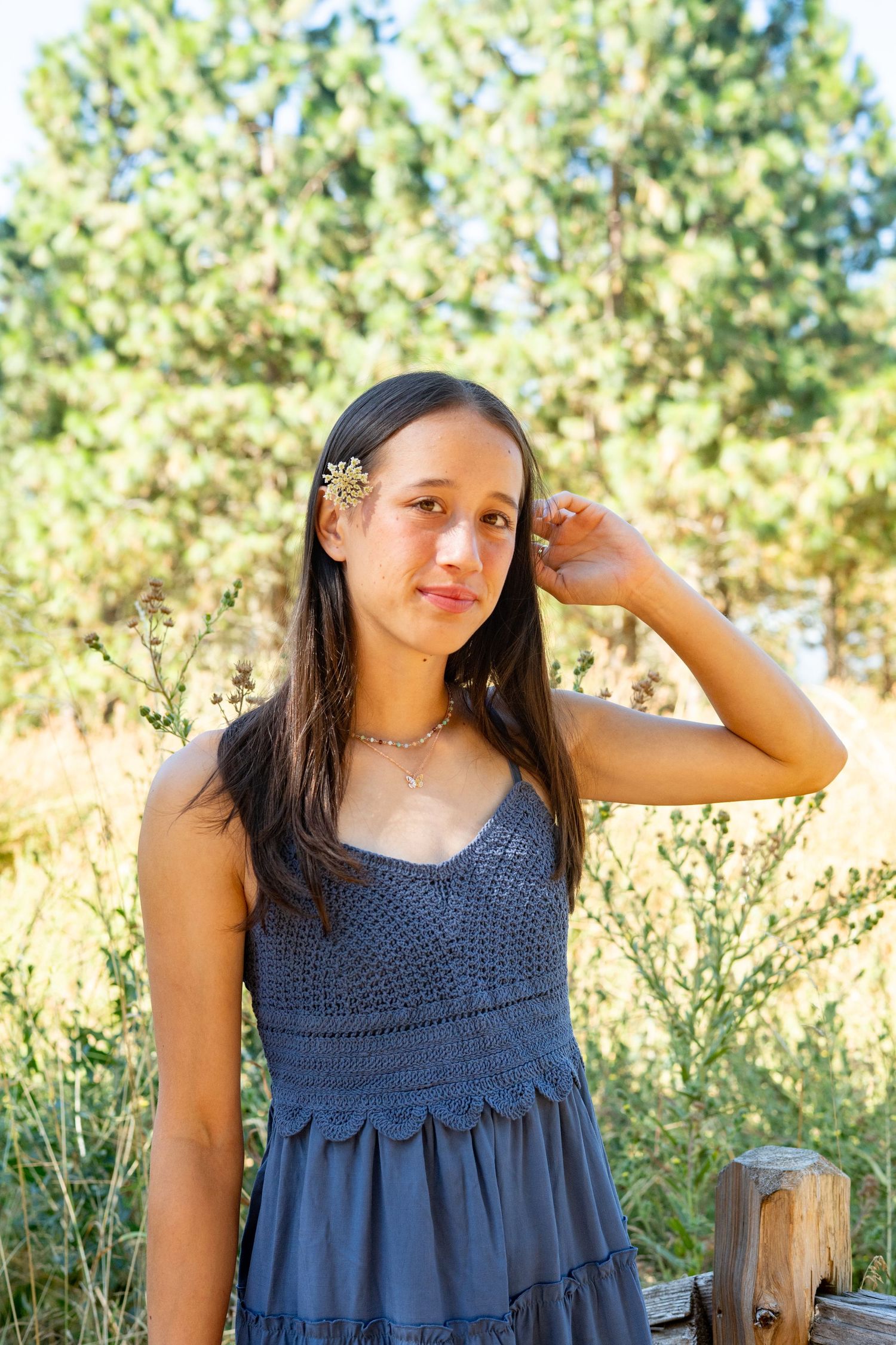 Young woman in navy blue summer dress poses outdoors with trees and foliage in background.