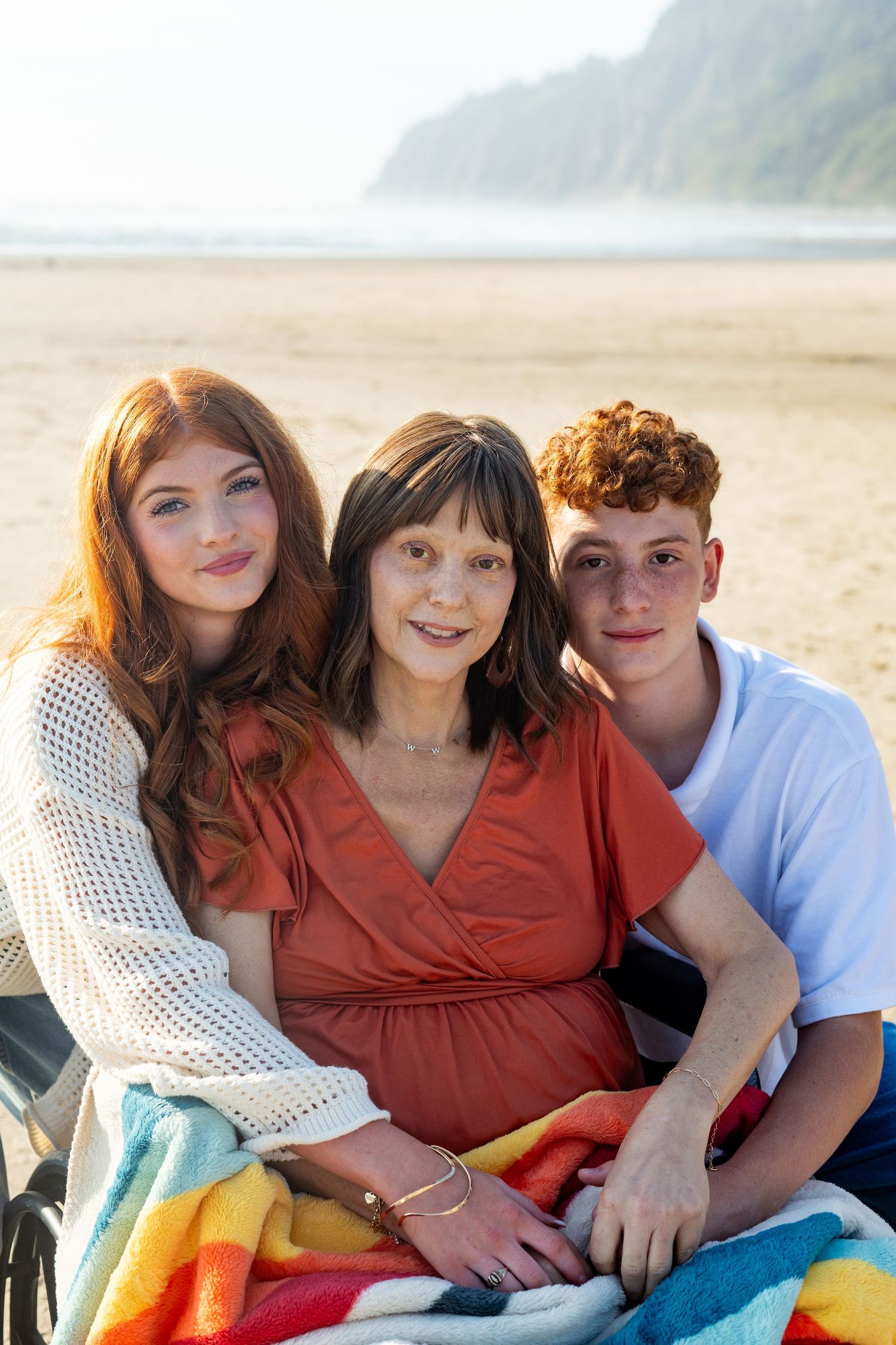 A close group sits together on a colorful beach blanket by the ocean on a sunny afternoon.