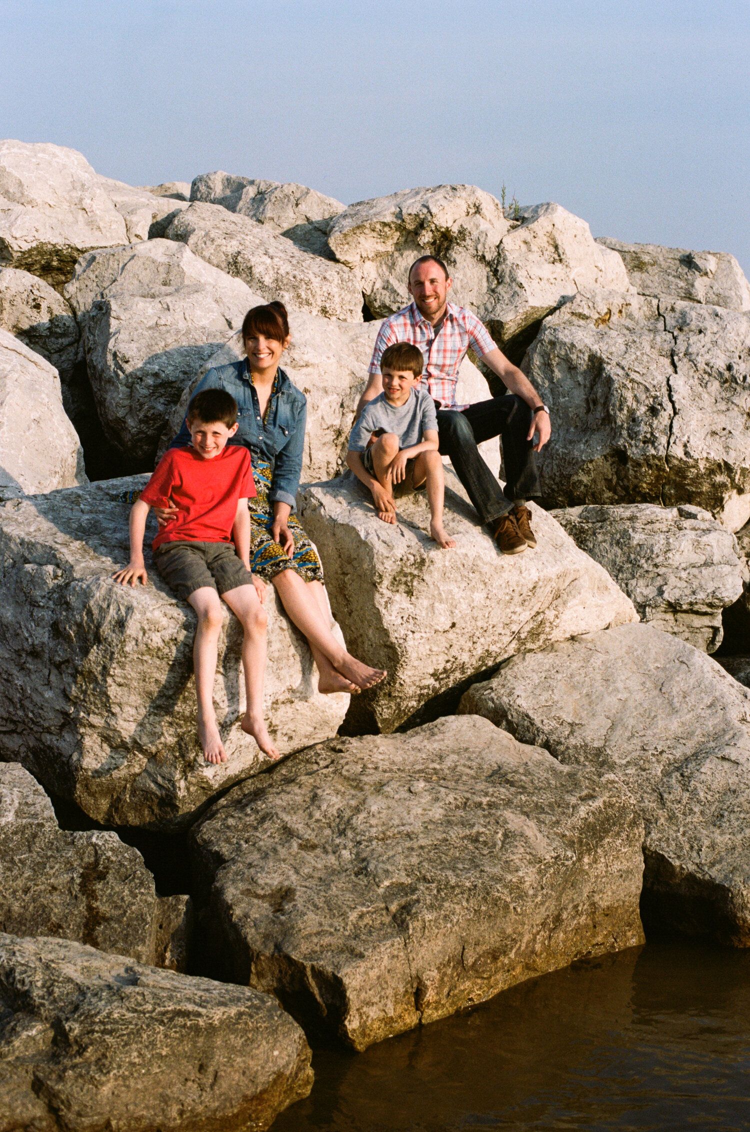 People relax on large coastal rocks next to calm waters during a sunny day.