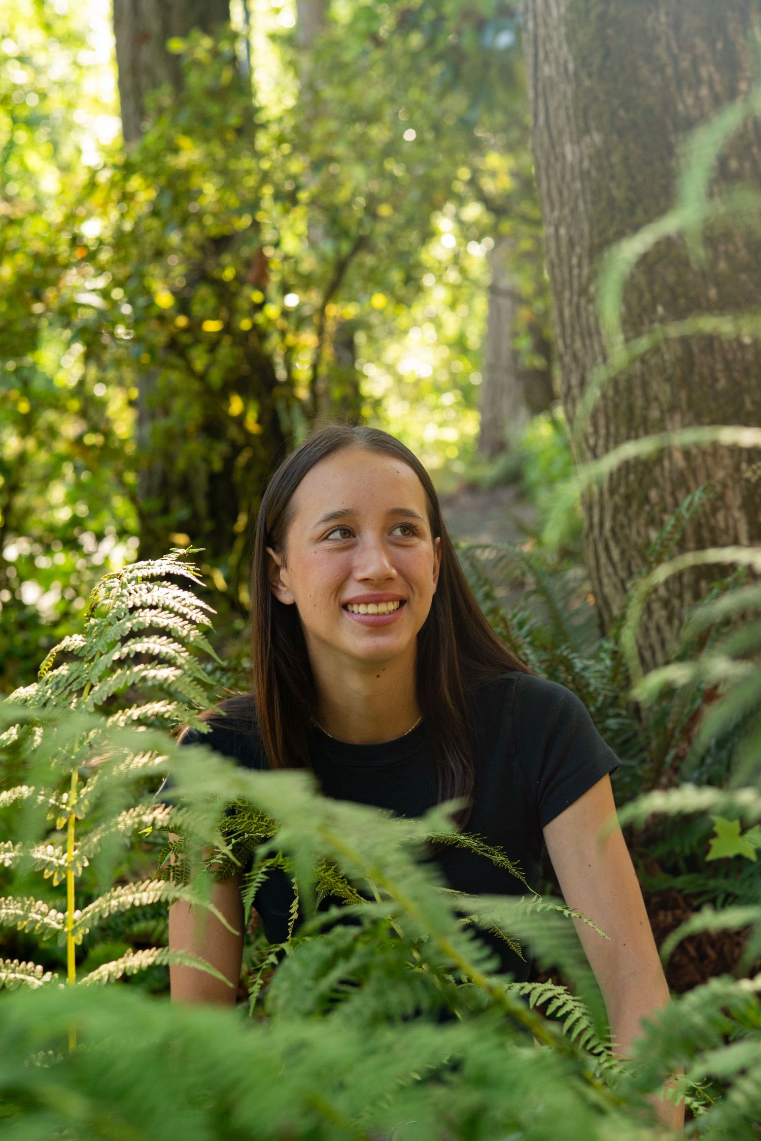 A person smiling while sitting among lush green ferns in a sunlit forest setting.