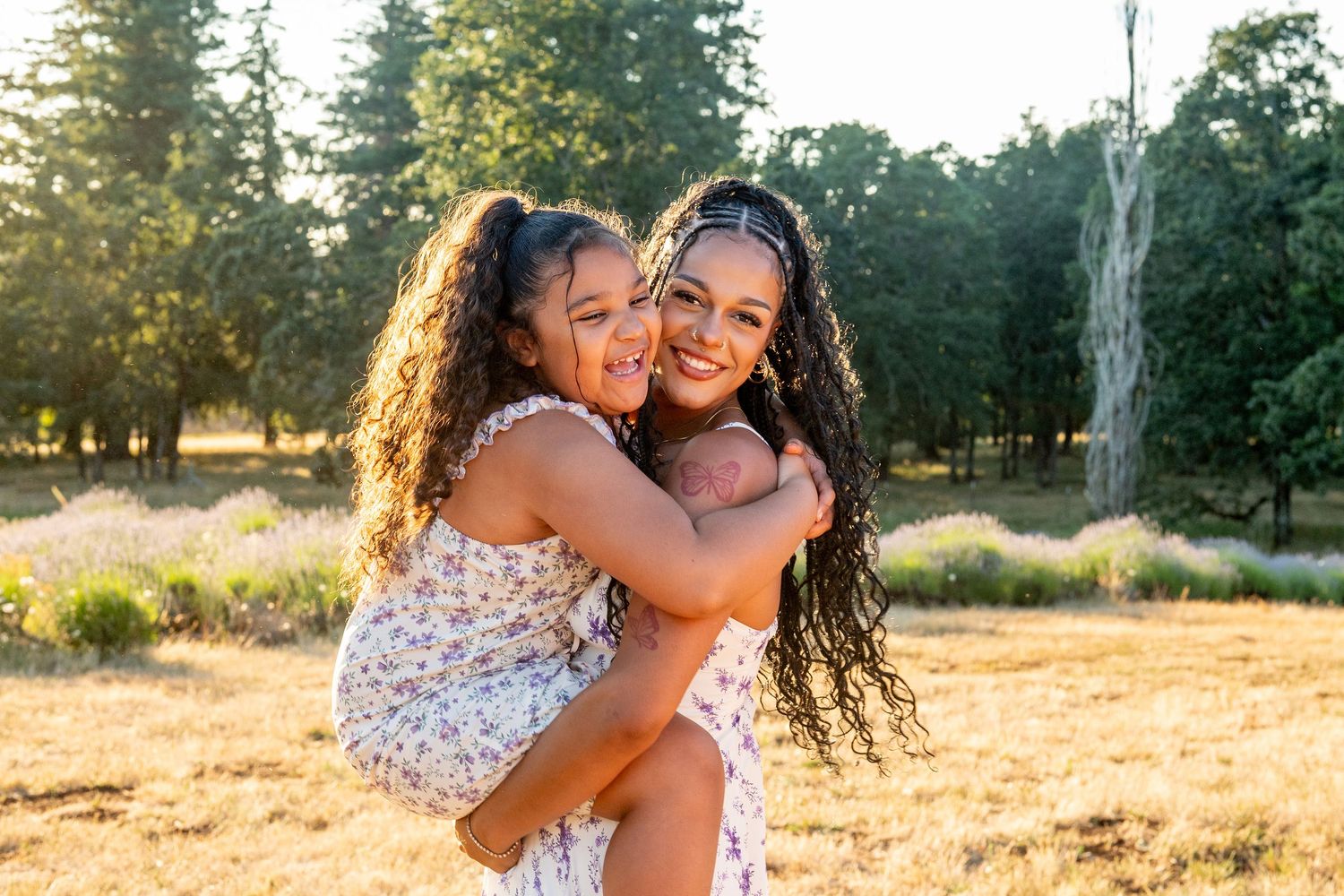 Two friends with curly hair sharing a joyful embrace outdoors in a sunny field at golden hour.