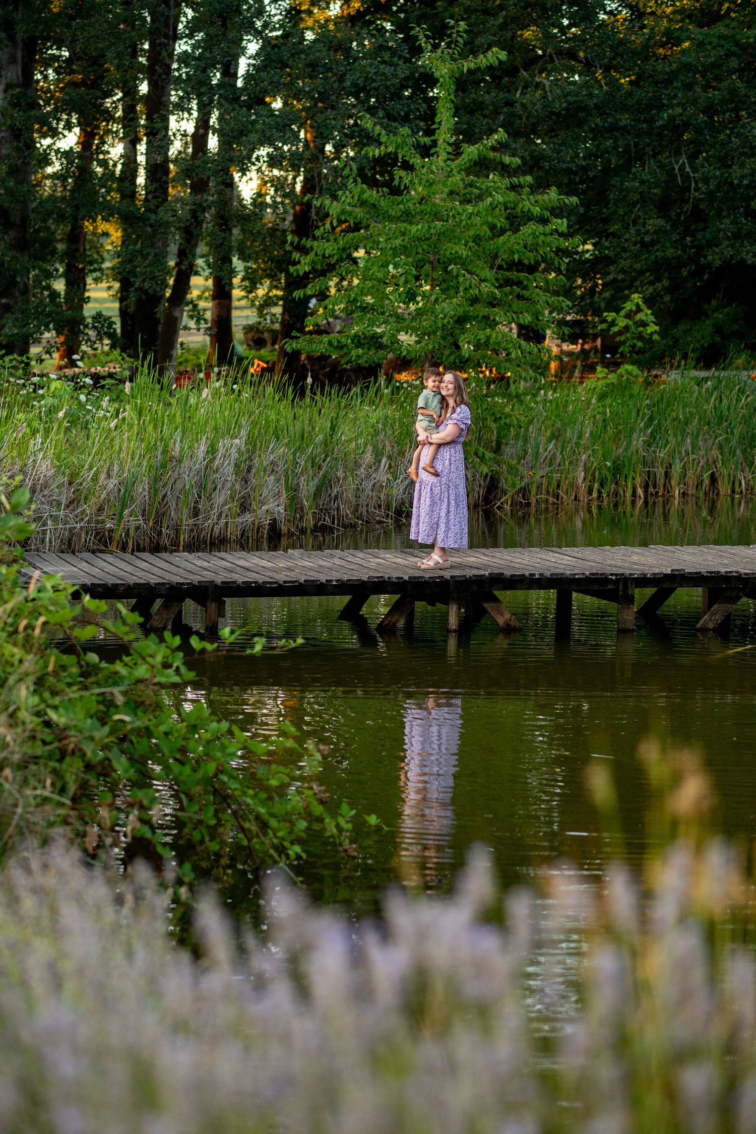 A couple stands on a wooden dock surrounded by tall reeds and trees reflected in a tranquil pond at sunset.