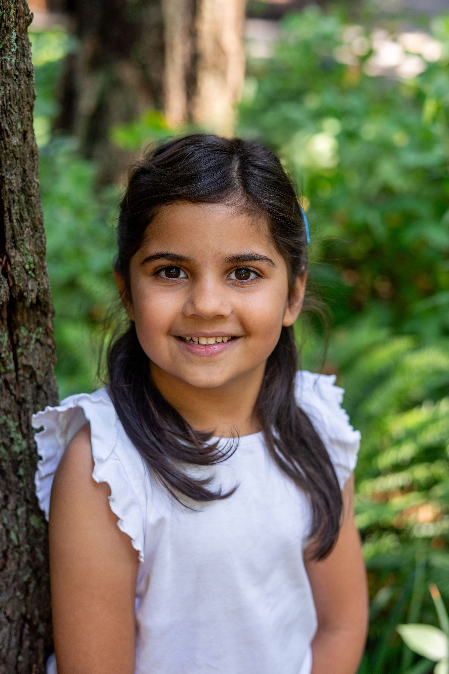 A smiling young child with dark hair and a white ruffled sleeve top poses against a blurry forest background.