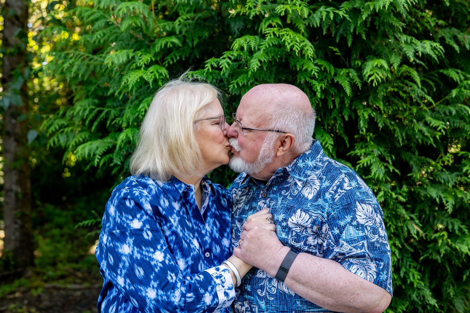 A couple in matching blue floral shirts stands in front of lush green foliage in a romantic outdoor setting.