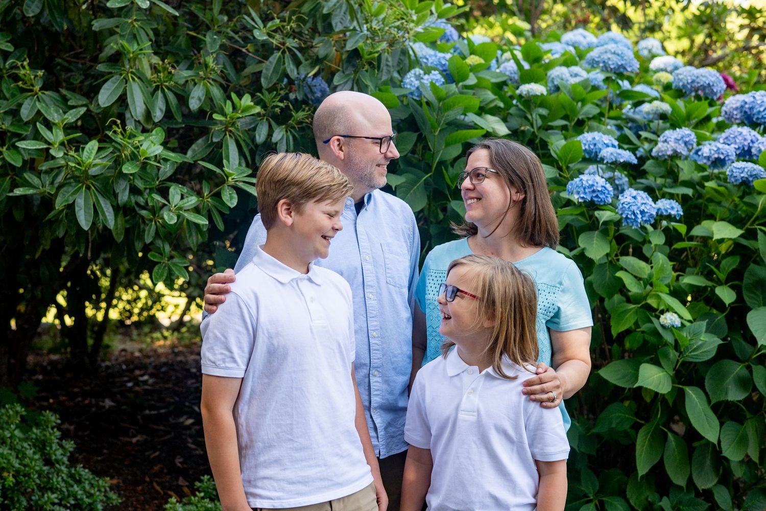 Family poses outdoors for portraits in a garden with blooming hydrangea bushes in the background.