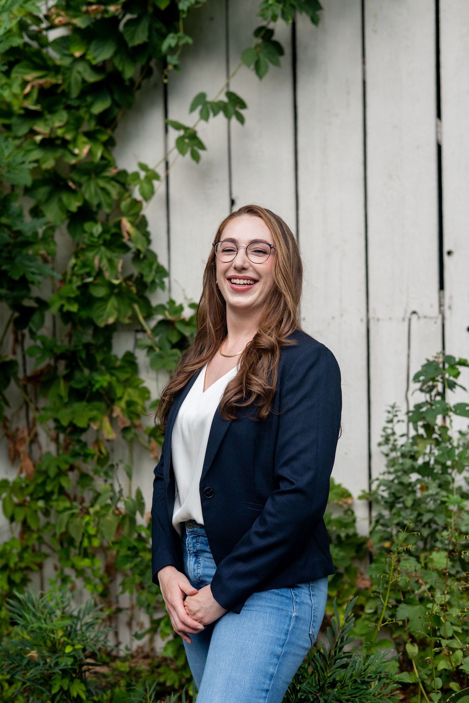A series of professional portraits taken against a white fence with green ivy, featuring glasses and a navy blazer.
