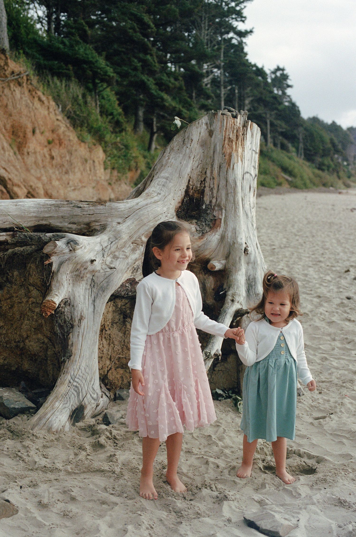 Two children in dresses stand by a large piece of driftwood on a sandy beach.