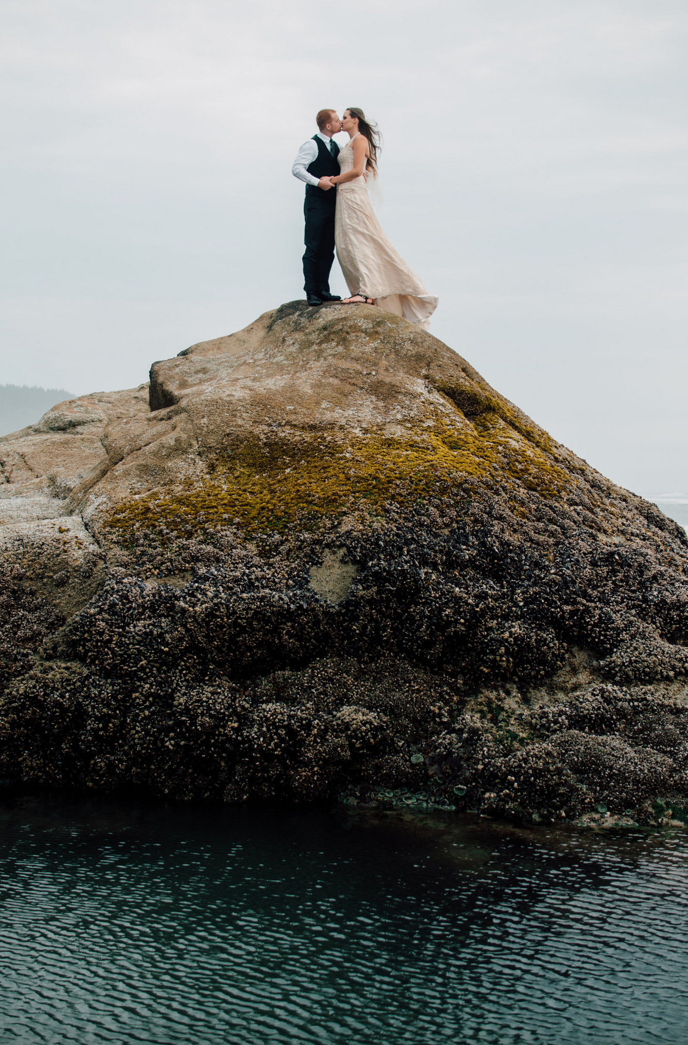 A couple in wedding attire embraces on top of a large coastal rock formation above turquoise waters.