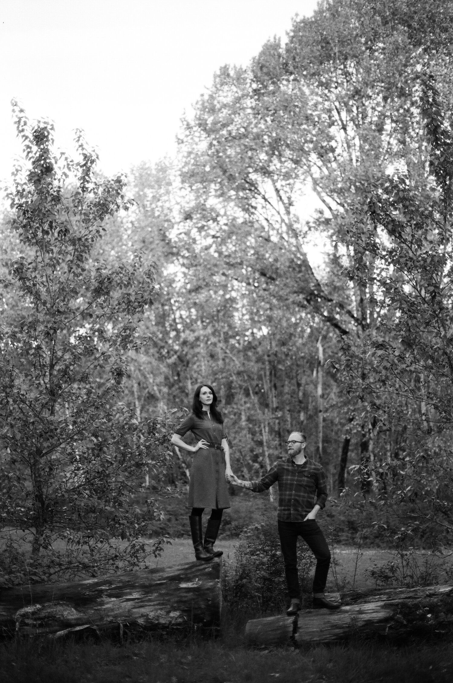 A black and white photograph of two people perched on fallen logs by a stream in a wooded forest setting.