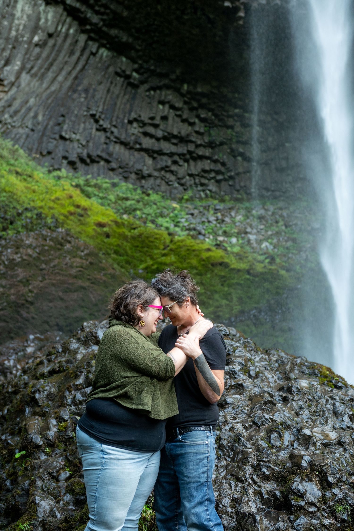 A couple shares a romantic moment in front of a majestic waterfall cascading down moss-covered rocks.